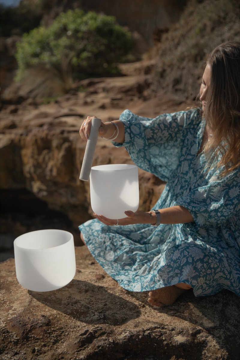 Kim Yates playing crystal singing bowls on the rocks by the ocean